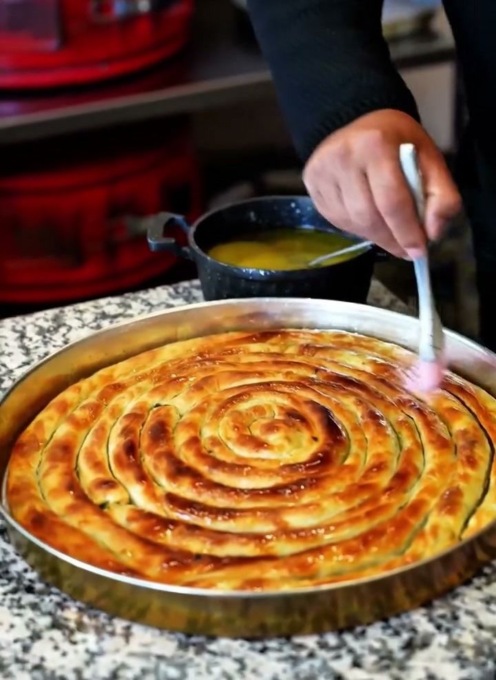 Fırından yeni çıkmış spiral börek tepsisine fırçayla tereyağı sürülüyor