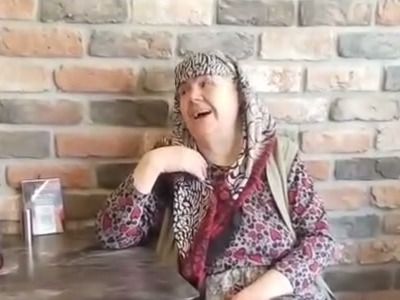 Furuna’da tuğla duvarın önünde oturmuş, kahkahayla gülen Naile teyze